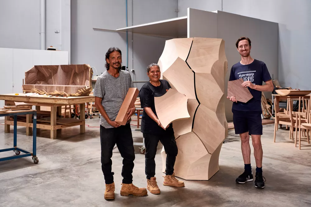 Three people stand smiling with a large-scaled furniture art piece, each holding different wooden elements.