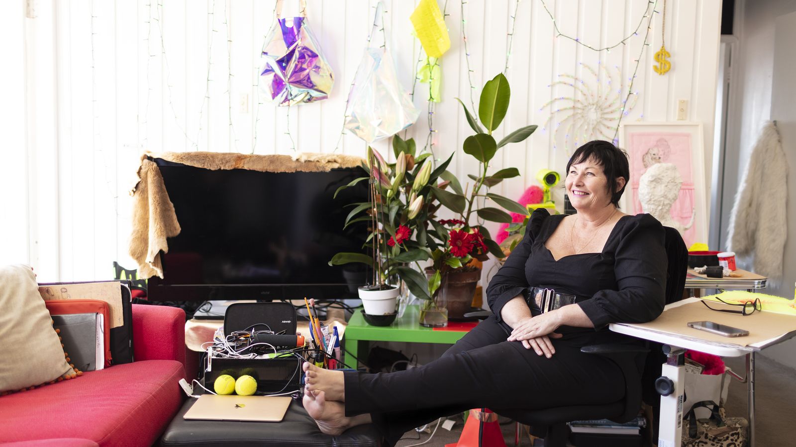 A women sits in her home studio dressed in black with plants, fairy lights and other objects.