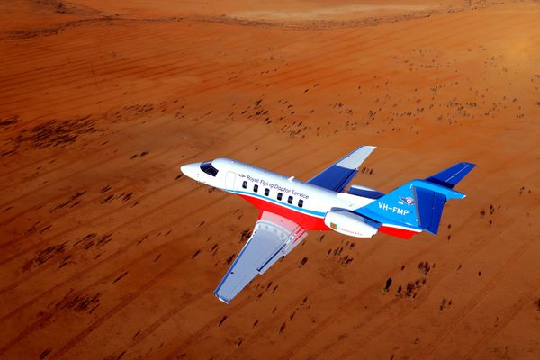 A medical jet aircraft with RFDS logo flies over a dry, brown landscape