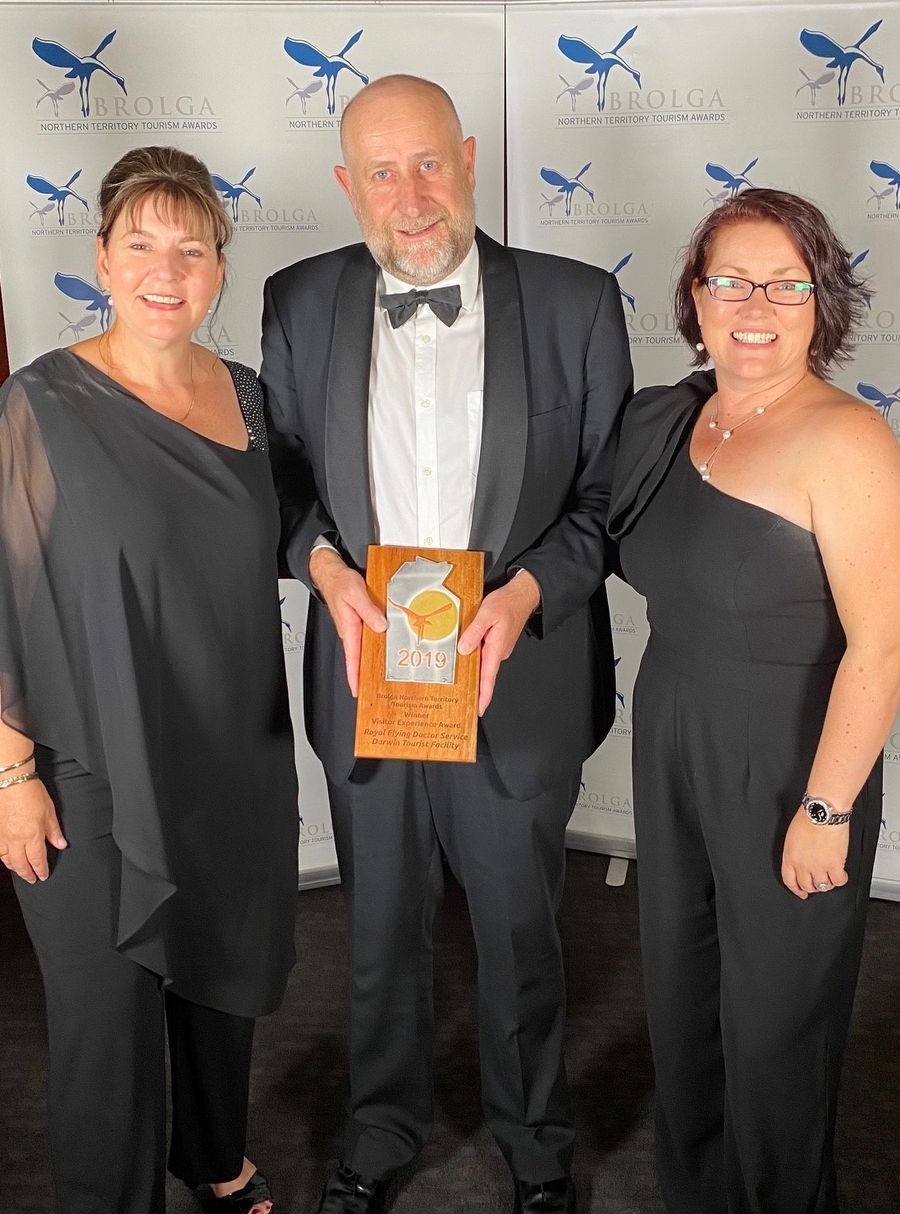 Three people in black formal wear smile at the camera. They are holding a trophy that reads 2019 BROLGA AWARD. 