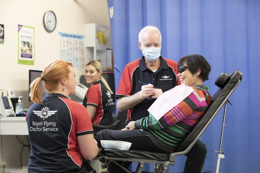 Four people sit in a medical office. A lady in a striped top sits in a dentist chair. A man wearing a dental mask sits next to her. 