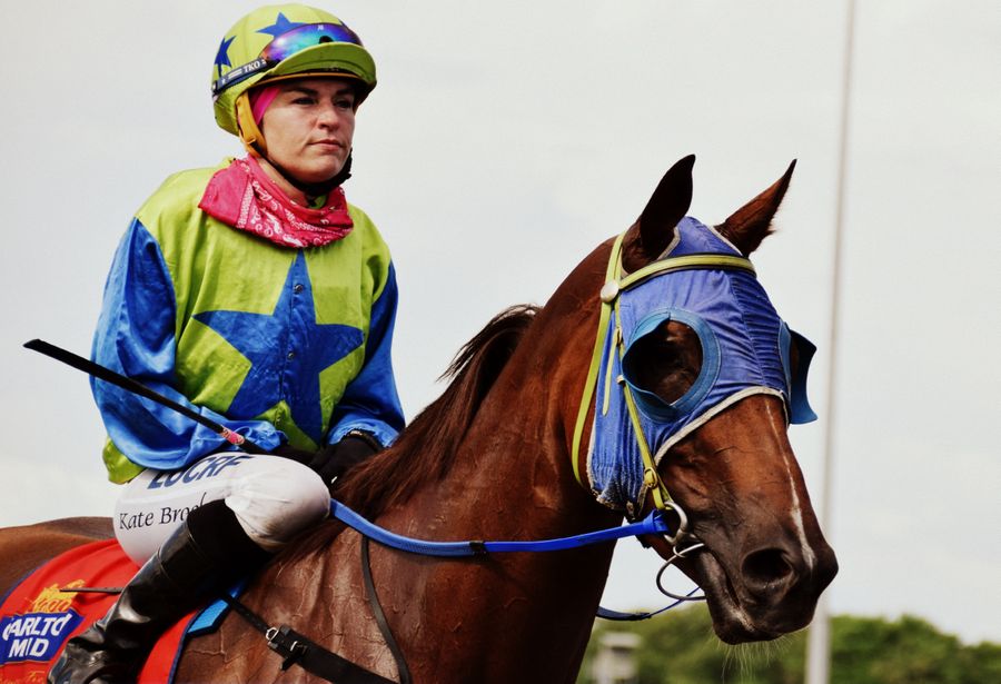 A young woman wearing horse racing silks and a brightly coloured helmet is sat atop a brown horse. 