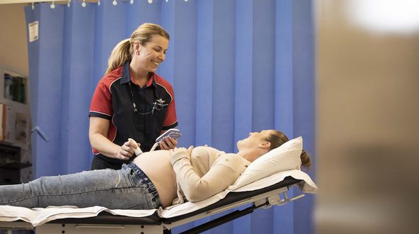 A pregnant woman lies on a hospital bed. A smiling female in a RFDS polo shirt holds a doppler over her pregnant tummy. They are both smiling.
