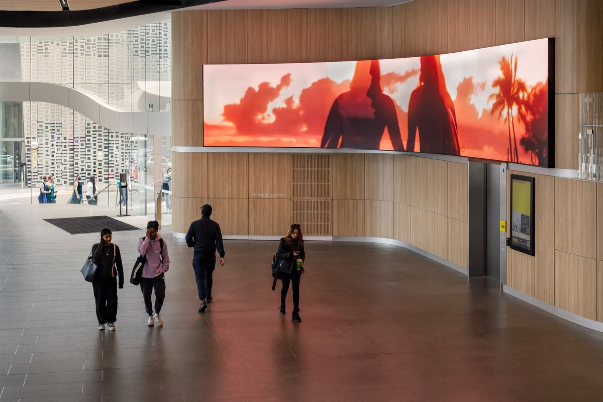 A large digital screen in a public space with a sunset and surfers walking on the beach
