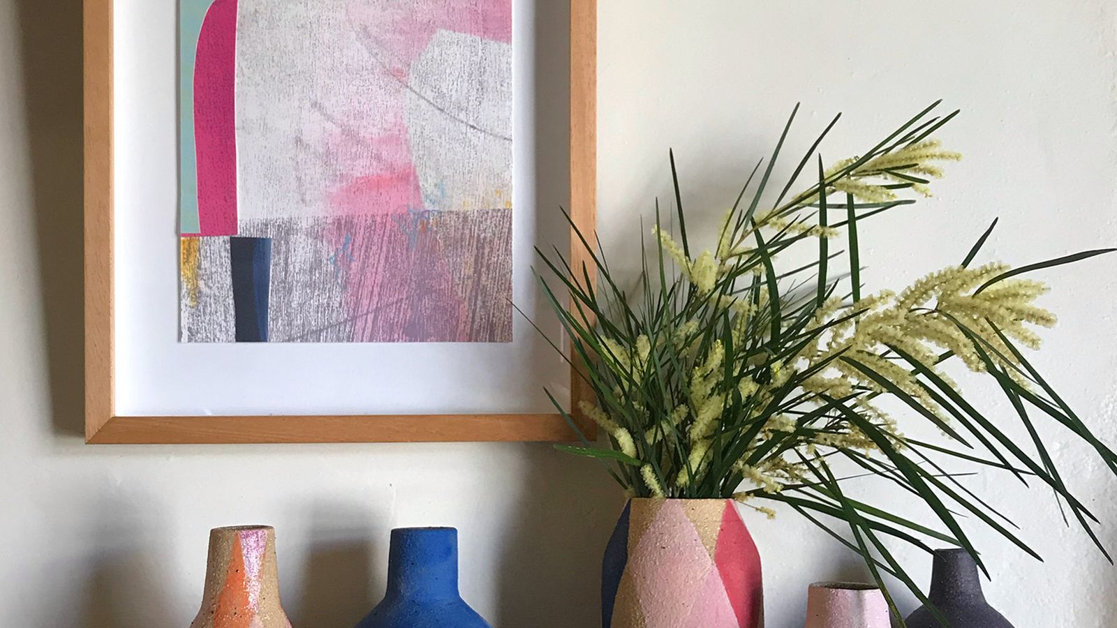 A group of brightly coloured ceramics vases sitting on a sideboard, below a bright coloured artwork.
