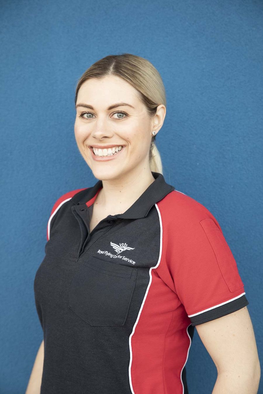 A woman with blonde hair smiles at the camera. She is wearing a red and navy polo shirt with RFDS logo on her chest. 