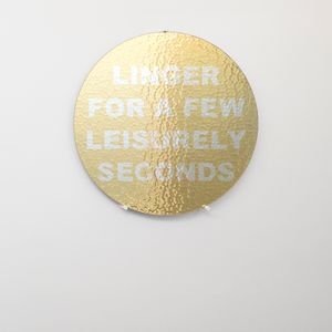 A big circular glass tabletop with bold white text on gold background that says "Linger For A Few Leisurely Seconds" setting on wooden a shelf against a white wall