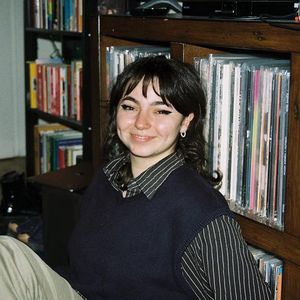 Lily sitting in front of a bookshelf, wearing a dark sweater with stripes on the sleeves and collar