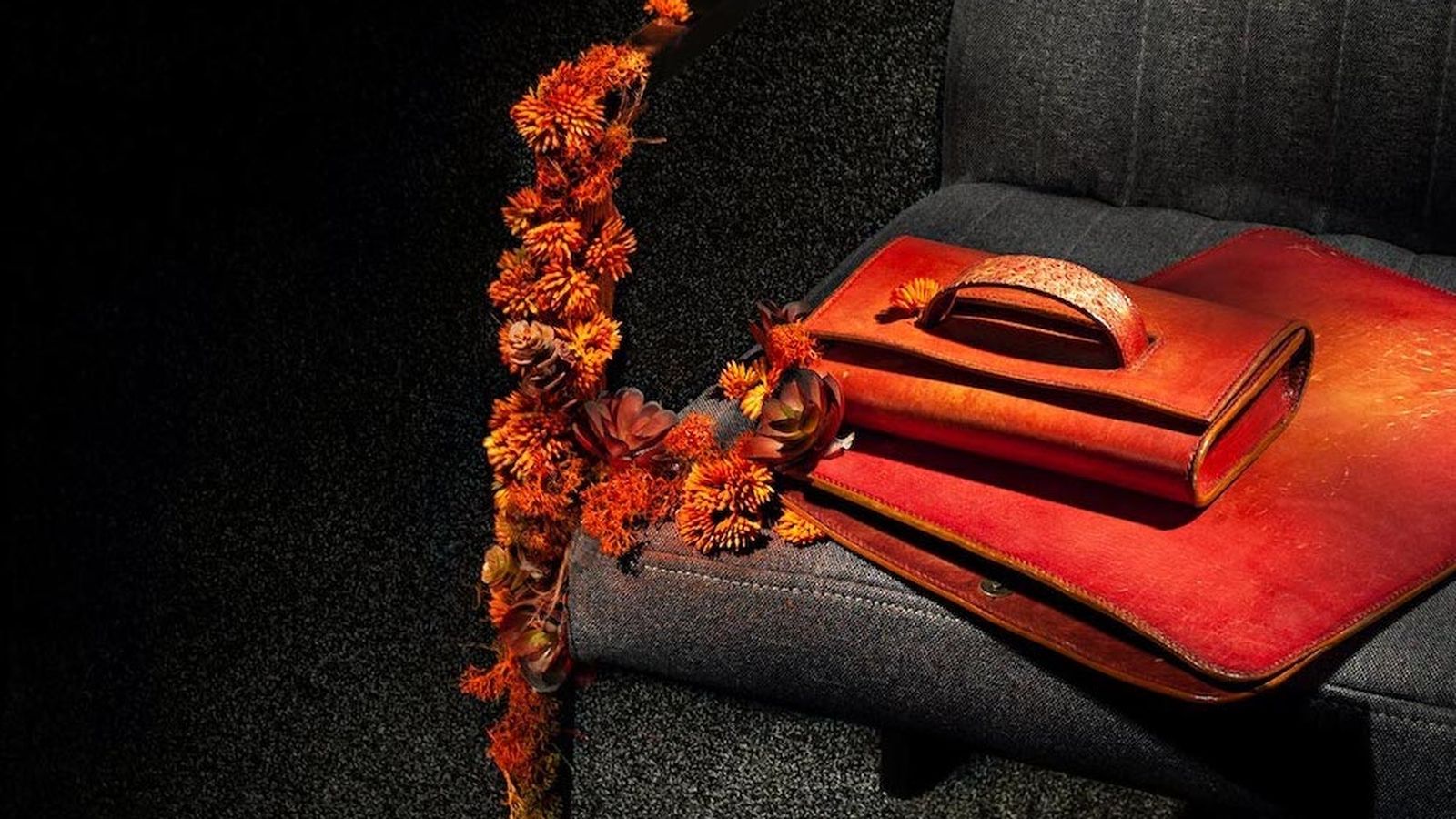 An orange leather handbag and satchel on a grey chair decorated with orange dried flowers