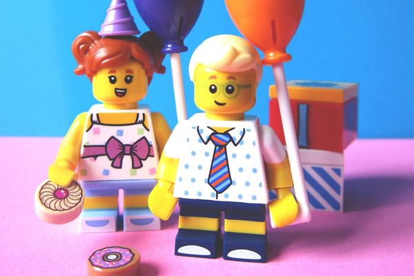 Two Lego mini-figures holding balloons and cake in front of wrapped presents