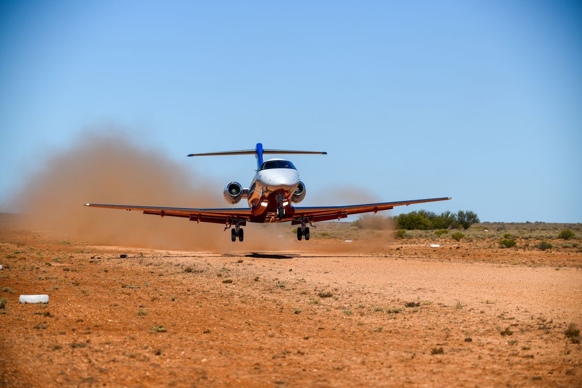 An aeromedical jet stands on tarmac. A woman in high vis vest pushes a stretcher with an adult patient lying on it toward the aircraft.