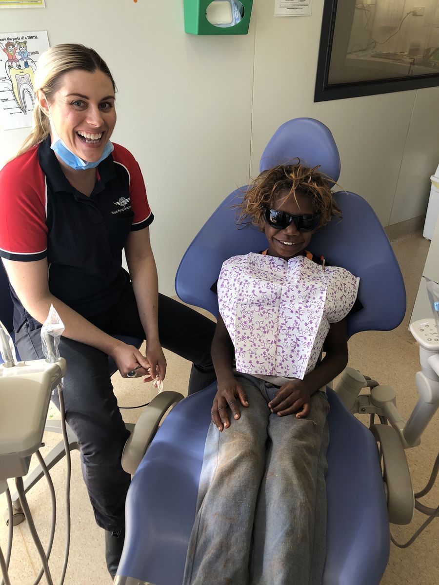 A woman wearing a RFDS shirt and dental masks sits next to a dentists chair. A young Aboriginal boy sits in the dentist chair. They are both smiling. 