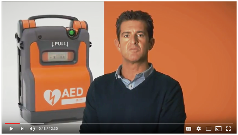 A still image from a YouTube video. The image features an orange AED and a man speaking. The background is orange. 