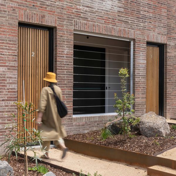 A section of a brick building facade with someone walking towards the wooden entrance door.