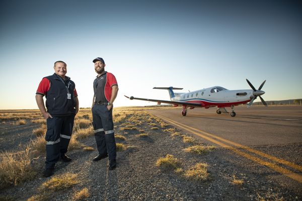 Two men in RFDS uniform stand smiling in front of a RFDS aircraft standing on a tarmac runway