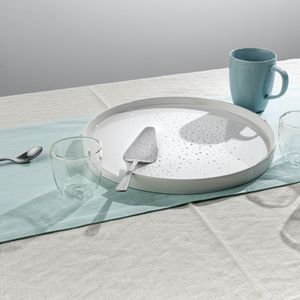 A white cake dish, metal cake server, teal coffee cup, metal spoon, and three glass cups arranged on a teal table runner