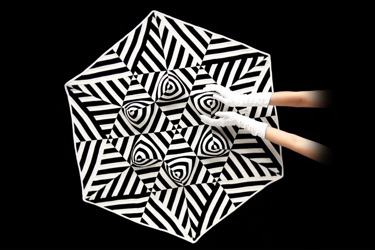 two arms and white gloved hands cupping part of the pattern created by the black and white illusion quilt in the background