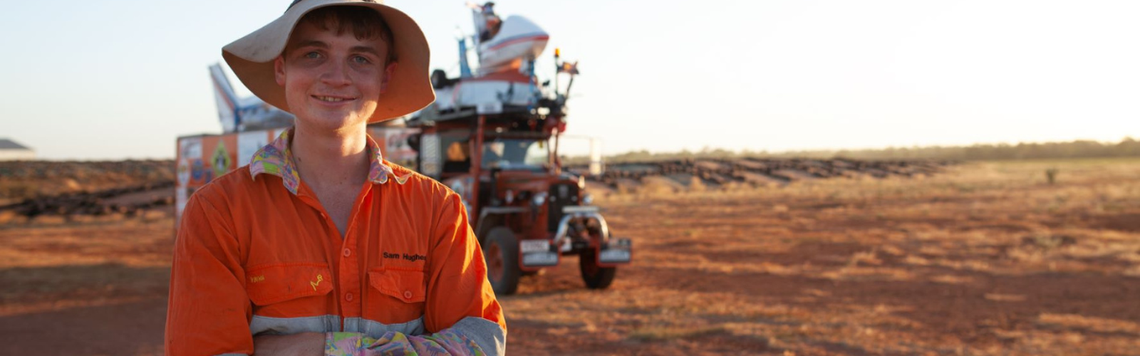 A young man stands in front of an orange tractor. He is a wearing bright orange work shirt and old hat and is smiling at the camera.