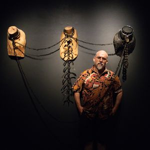 A portrait image of Dominic White with a serious expression wearing a brown patterned shirt and standing in front of his artwork, Collared, installed on a black wall..