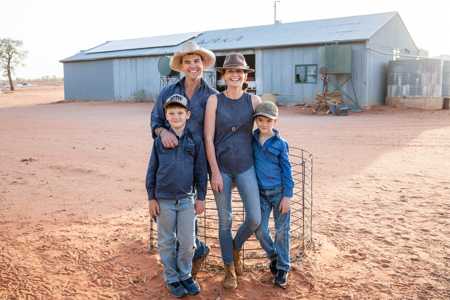 Family of four standing in front large shed on outback farm. 