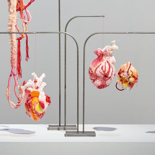 A wide shot of the exhibition Hearts of Absent Women showing two metal stands with hanging embroidered hearts in pinks, reds, yellows and oranges and a wall tapestry of a pelvis with flowers growing out of it.