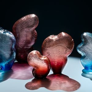 5 pieces of cloud or puffs of air shaped blown and carved glass in blue, red and purple sit on a gray top and cast shadows