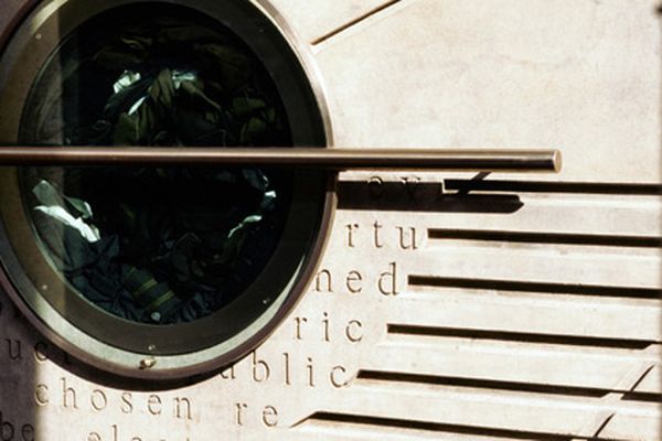 Photo of a concrete wall with horizontal indents in it and a circular window inset to the left of the image with clothing inside.