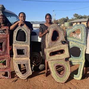 Photo of four women holding up woven sculptures on a red dirt road in Central Australia