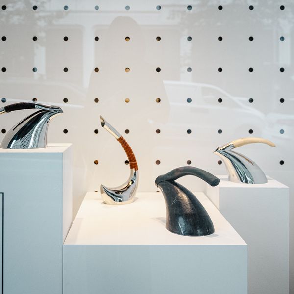 Image of four stainless steel sculptural kettles with various handles, celery top pine to kangaroo leather, arranged on white plinths white white peg board background