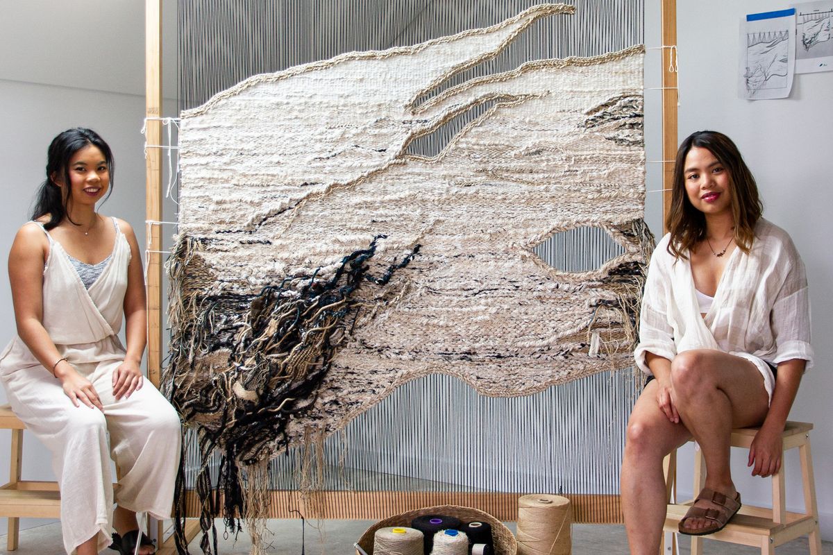 Two women sit on small timber stools, between them is a large textile work attached to a loom