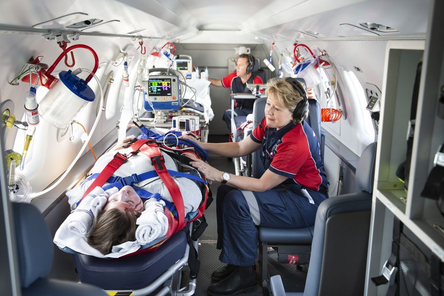 Interior view of aeromedical jet. Two medical staff in uniforms tend to a patient lying on a hospital stretcher. Her head is bloody. 