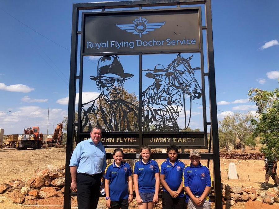 A man stands with a group of schoolchildren in an outback Australian scene. There is a large iron sign that reads RFDS. 