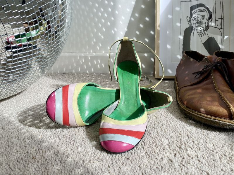 A pair of colourful high heels, A pair of brown leather boots, a disco ball and a framed drawing are grouped together on the floor