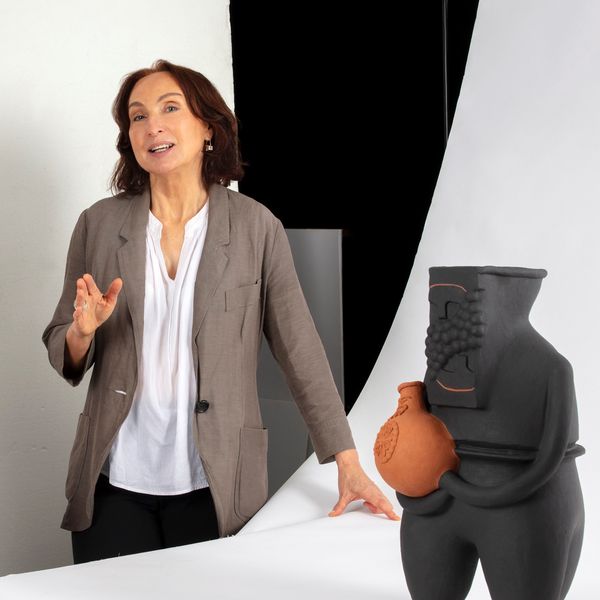Person with shoulder length brown hair wearing greige blaze talking to camera with a ceramic figure in foreground.