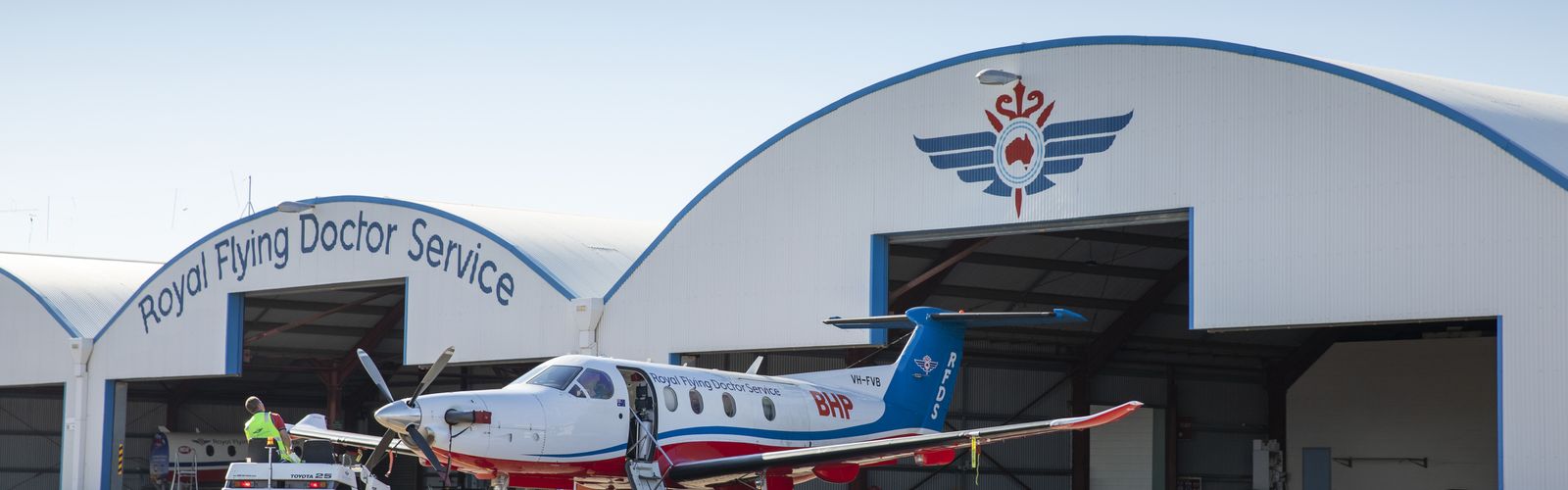 Two men in RFDS uniform stand smiling in front of a RFDS aircraft standing on a tarmac runway