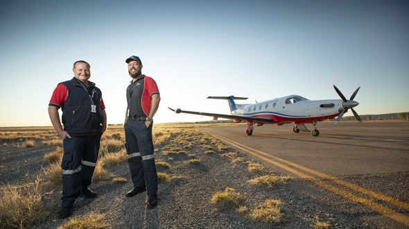 Two men in RFDS uniform stand smiling in front of a RFDS aircraft standing on a tarmac runway