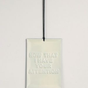 A rectangular silver sign with the text "NOW THAT I HAVE YOUR ATTENTION" hanging on a black cord against a white wall