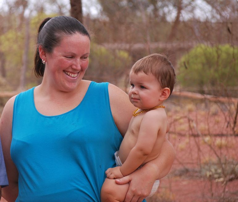 A woman in a blue singlet holds a baby in a nappy. She is smiling. 