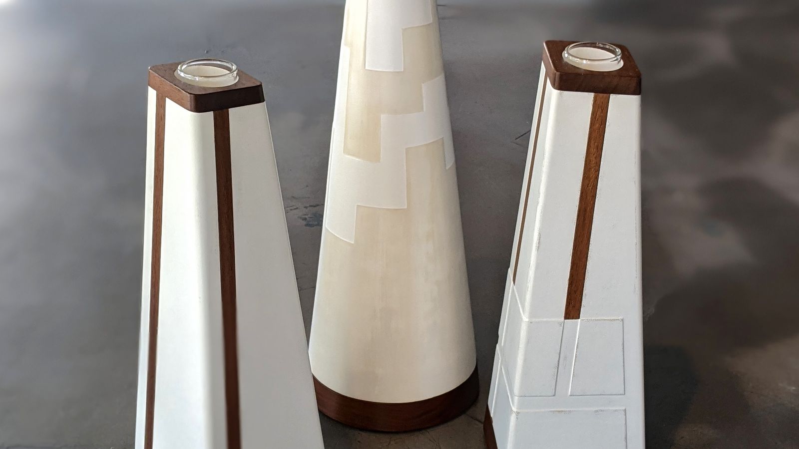 Three tall pyramid shaped vases with white and wood finish standing on grey floor