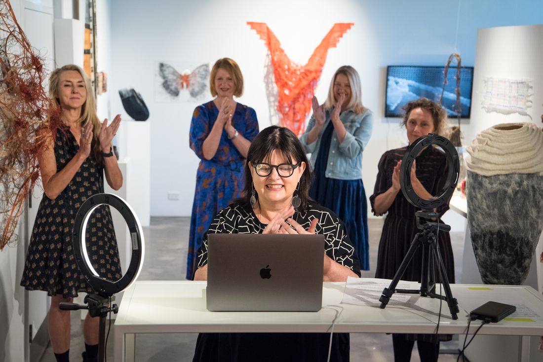 Photo of a woman with lights speaking into a computer for a zoom opening with four women standing  behind in a gallery space.