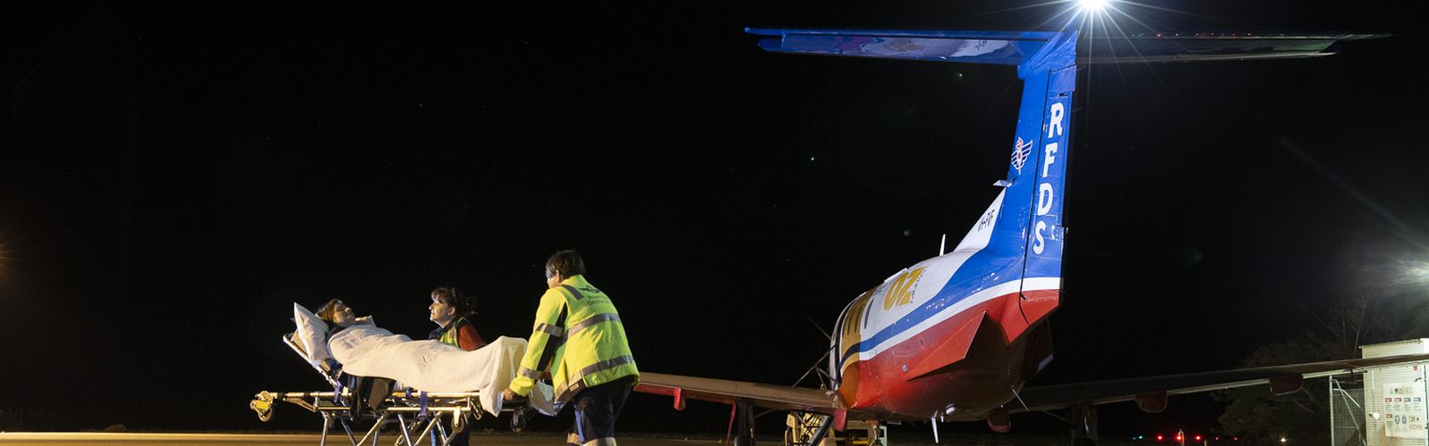 An aircraft with RFDS logo flies over green paddocks. 