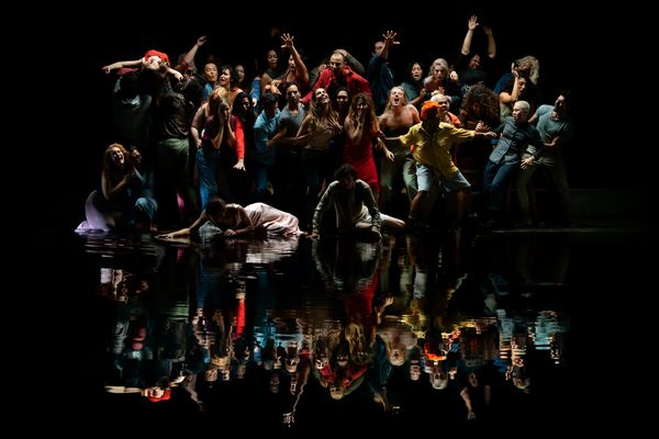 A crowd of people surge around a central figure who is seated, gazing at their own reflection in dark water.