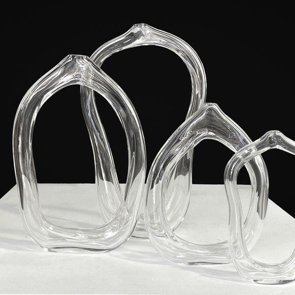 Four transparent glass sculptures are showcased in this image. Each sculpture resembles an abstract, distorted form of a heart or loop. They are elegantly placed on a white surface against a dark background, creating a striking contrast.