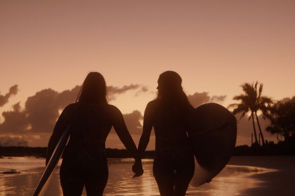 An image of two women in silhouette against a sunset walking with surfboards on the beach.