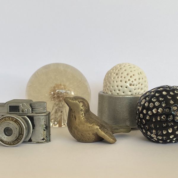 A group of objects, including a small vintage camera, a brass bird, a black ceramic ball, a white ceramic bowl, a metal dish and a paperweight made from a dandelion encased in resin