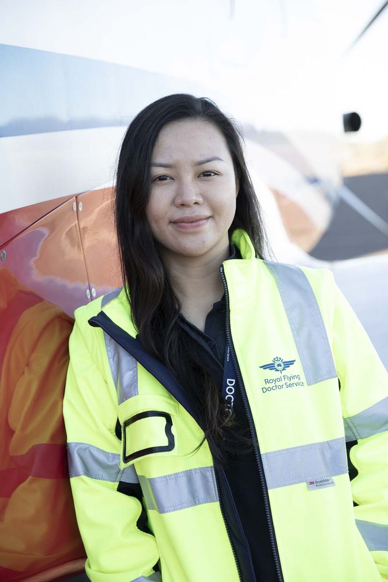 A woman with long dark hair smiles at the camera. She is wearing a yellow high-vis jacket and a lanyard that says DOCTOR. 