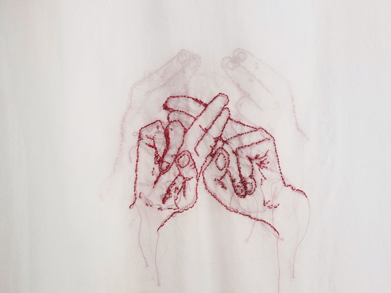 Two hands forming the ASLAN sign for family stitched in red on white fabric