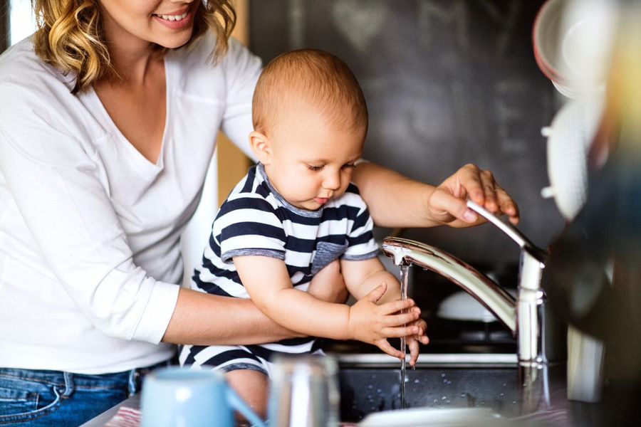 Mother holding a baby while playing with a kitchen sink tap