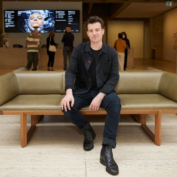 Photo of a person with short brown hair wearing black pants and jacket looking to the camera and sitting on a wooden bench in a marble foyer.