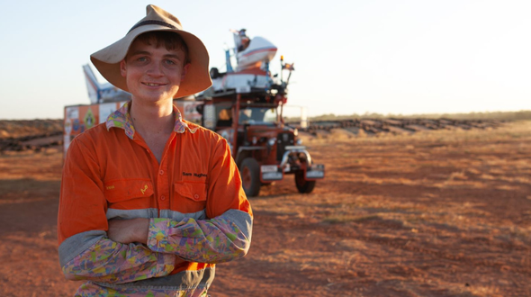 A young man stands in front of an orange tractor. He is a wearing bright orange work shirt and old hat and is smiling at the camera.